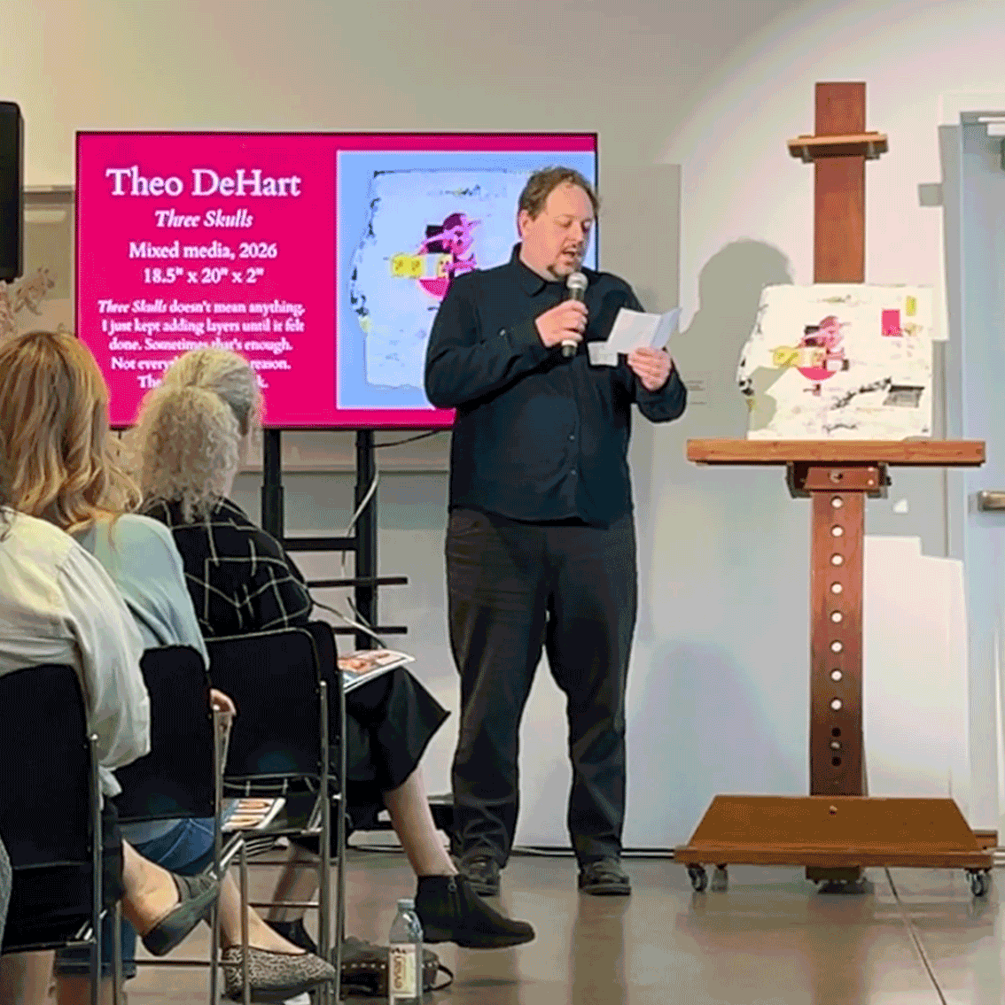 Theo DeHart stands at a microphone reading from a paper at a critique event. Behind him a pink slide reads "Three Skulls, Mixed media, 2026, 18.5 x 20 x 2 inches." The piece sits on an easel to his right. An audience watches from the foreground.
